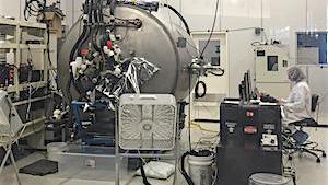 Looking inside a clean room with a large metal circular chamber with an individual wearing  a cleanroom suit sits at a computer nearby.