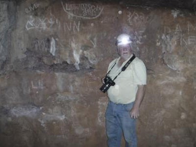 An adult male stands next to a cave wall wearing a helmet and a camera around his neck.