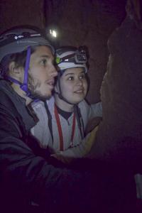 Two students wearing helmets with lights examine a cave wall.