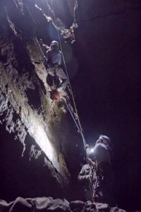 Two unidentifiable people descend down a cave wall, while a third person holds the rope from the bottom.
