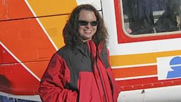Angie stands in a red coat, hands in her pockets, in front of a helicopter.