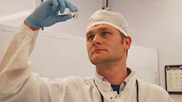 Greg wearing a white lab coat, head covering and gloves, inspects a specimen he holds in his hand.