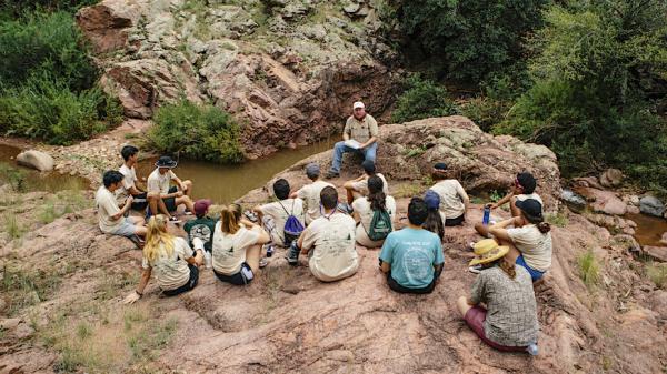 A group of students sit together on the rocks listening to a leader as he sits above.