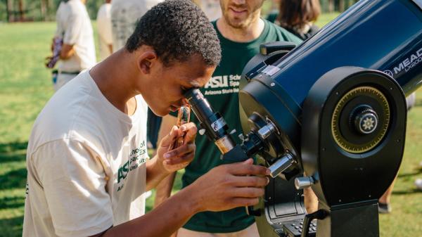 A daytime scene of a  male student looking through a telescope while another male stands nearby.