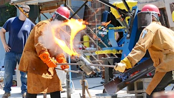 Two men in fire retardant suits and equipment, work outdoors with a large flame.