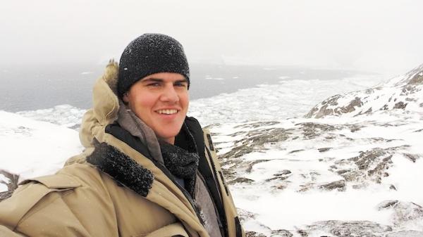 Jim smiles while wearing a heavy coat and hat in a snow-covered Greenland scenery.