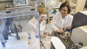 Two female researchers in white lab coats and goggles working in a lab.