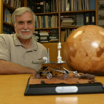 Ronald Greeley sits casually at a desk, a globe at his side and a model of a rover in front and a full book shelf behind him.