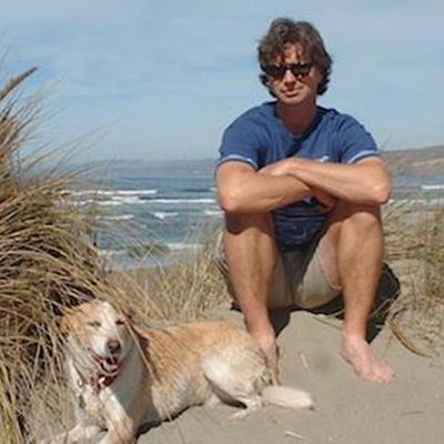 Jeff sits on a sandy beach with his dog, the surf to his back.